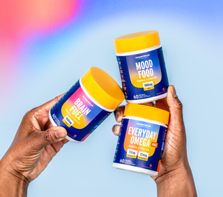 Two hands holding three supplement bottles against a colorful background. The bottles are labeled Brain Fuel, Mood Food, and Everyday Omega, each with yellow lids and designed for cognitive and mood support.