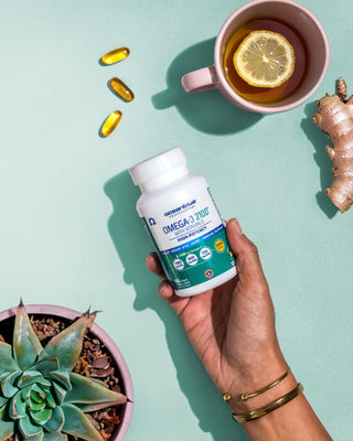 A hand holds a bottle of Omega-3 supplements next to a pink cup of tea with a slice of lemon. Near the cup is a piece of ginger. Three capsules are placed above, and a small potted succulent plant is seen on the left. The background is mint green.