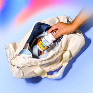 A hand places a bottle of vitamins into an open beige gym bag. The bag contains various items, including a water bottle and sneakers, against a colorful, blurred background.