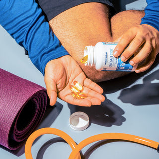 A person sitting cross-legged on the floor is pouring omega-3 fish oil capsules from a bottle into their hand. A rolled-up purple yoga mat and an orange resistance band are nearby on the light blue surface.