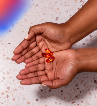 Two hands are cupped together, holding four glossy red capsules. The background is a light-colored terrazzo surface with blue and red light reflections on the left side.