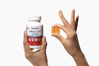 A hand holding a white bottle labeled Omega-3 2100 and another hand holding two orange capsules against a white background. The bottle features various symbols indicating its benefits and potency.