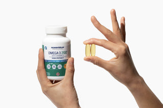 A hand holds a bottle of Oceanblue Omega-3 2100 with Vitamin D supplements. Another hand holds two large yellow tablets between fingers. The background is plain white.