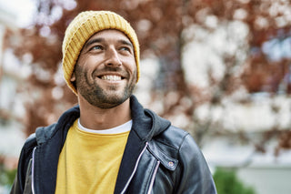 A man wearing a yellow beanie, a yellow sweater, and a black leather jacket smiles while looking off to the side. The background features blurry autumn trees with red leaves.