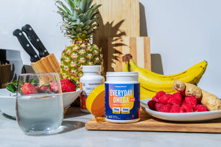 A countertop with a glass of water, strawberries, and raspberry, banana, ginger slices, and pineapple. Omega supplements are in focus in the center. Knives in a block and a wooden cutting board are in the background.