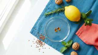Professional flat lay of amber omega-3 softgels with lemon, walnuts, flaxseeds, and herbs on an ocean-blue surface