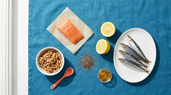 Flat-lay arrangement of omega-3 rich foods including salmon, sardines, walnuts, chia seeds, lemons, and amber softgels on an ocean-blue linen