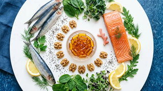 Flat lay of amber softgels, salmon, walnuts and leafy greens on a bright white surface