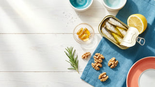 Flat lay of sardines, amber omega-3 softgels, lemon, walnuts, and rosemary on a white coastal table