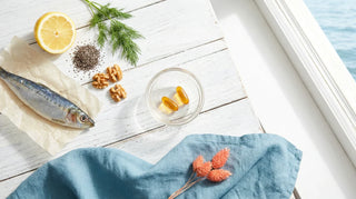 Flat lay of amber omega-3 softgels, sardine, lemon, chia seeds, and walnuts on a bright coastal-themed white surface
