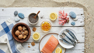 Flat lay of salmon, sardines, walnuts, chia, lemon, and amber softgels on white driftwood by the coast