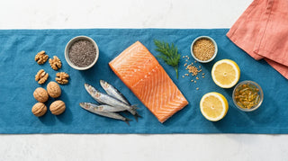 Flat lay of salmon, sardines, walnuts, chia, flax, lemon and amber softgels on white with blue linen