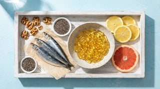 Flat lay of fish oil softgels, sardines, walnuts, chia seeds, and lemon on a bright white coastal-themed surface