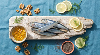 Flat lay of sardines, omega-3 softgels, walnuts, chia seeds, and lemon on a driftwood board with ocean-blue linen