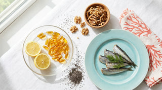 Flat lay of amber omega-3 softgels, sardines, walnuts, chia seeds, and lemon on a bright white linen surface with ocean-blue accents