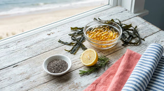 Flat lay of amber omega-3 softgels in a glass dish with chia seeds, lemon, and rosemary on a driftwood surface near a coastal window