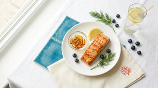 Flat lay of grilled salmon, omega-3 softgels, blueberries, and lemon water on a bright white surface