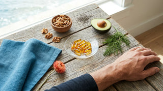 Flat lay of amber omega-3 softgels, walnuts, avocado, and chia seeds on a coastal-style driftwood table