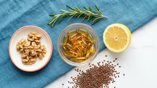 Flat lay of amber omega-3 fish oil softgels in a glass bowl with lemon, walnuts, flaxseeds, and rosemary on an ocean-blue linen background