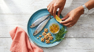 Flat lay of sardines, amber omega-3 softgels, walnuts, and fresh dill on a white wooden surface with ocean-blue plate