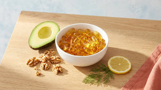 Flat lay of amber omega-3 softgels with avocado, walnuts, lemon, and dill on a light wood surface in natural light