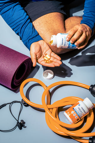 Person sitting on a floor holding vitamin capsules from a bottle in one hand. Nearby are a purple yoga mat, orange resistance band, and earphones.