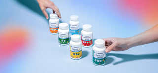 Two hands reach for white bottles on a blue surface. The bottles are labeled Omega 3 and feature colorful labels in orange, blue, and green, displaying supplement information. Soft blurred colors frame the image corners.