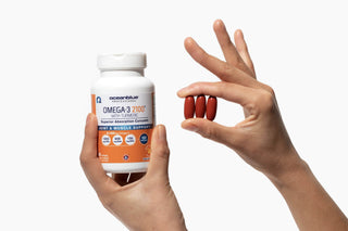 A person holds a bottle of Omega-3 2100 mg supplements with turmeric in one hand and three red capsules between their fingers in the other hand. The label highlights joint and muscle support features.