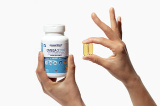A person holds a bottle of Oceanblue Omega-3 2100 supplements with Vitamin K2 against a white background. Another hand shows two yellow capsules next to the bottle, highlighting the pill size.