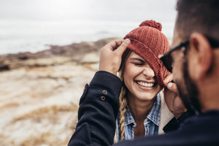 A person playfully pulls down a woman’s red beanie over her eyes, making her laugh. They are outdoors by a rocky landscape with the sea in the background. The woman is wearing a denim jacket and has long braided hair.