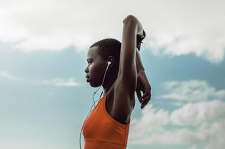 A person in an orange top stretches their arms overhead against a backdrop of a cloudy sky. They are wearing earphones and appear to be preparing for exercise.