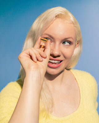A woman with long blonde hair is playfully holding a yellow ring up to her eye, smiling. She is wearing a yellow top against a bright blue background.